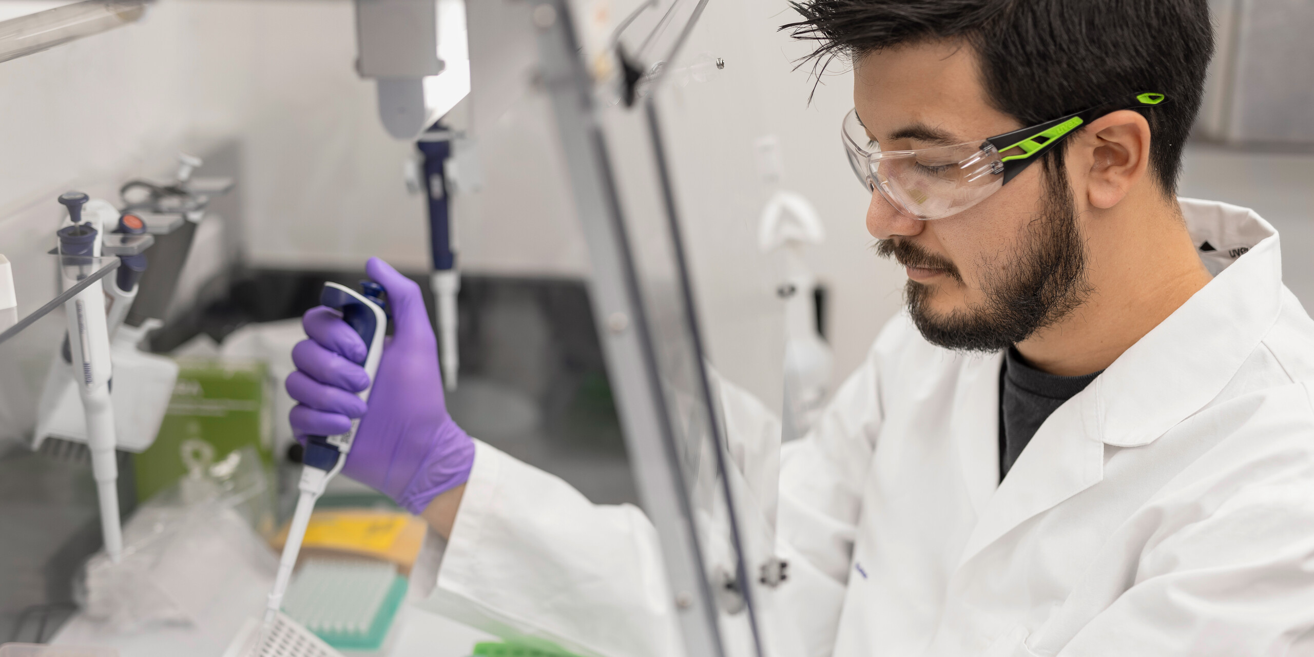 Male in lab attire pipetting material under a hood.