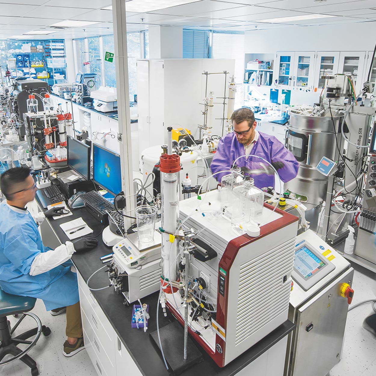 Two people wearing lab attire working in front of bioreactors.